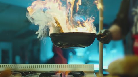 Professional chef cook flambe style . Prepares dish meat in a pan with open flames . Chef frying mixing food on gas hob in commercial kitchen . Man frying steak in flaming pan on hob in restaurant .  