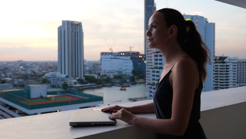 Evening hour, freelancer woman shut down laptop and look to city. She stand at balcony on high floor, blurred Bangkok city buildings on background, dim natural light | Shutterstock HD Video #1076758265