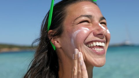Close up of happy smiling young woman is applying sunscreen or sun tanning lotion on face to take care of her skin on seaside beach during holidays vacation trip. Concept of healthcare and cosmetics.