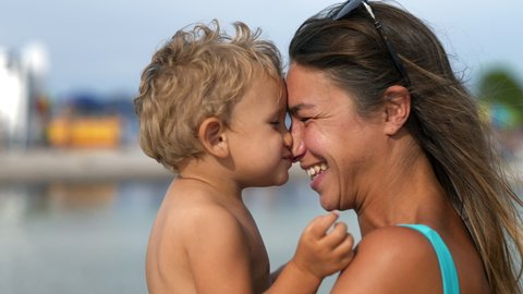 Authentic shot of young happy smiling mother is keeping on her arms and having fun to play nose to nose with her toddler baby boy on seaside beach in sunny day during family holidays vacation trip.