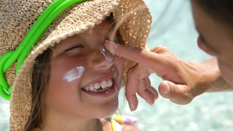 Close up of young happy mother applying protective sunscreen or sunblock lotion on her little happy smiling daughter's face to take care of skin on seaside beach during family holidays vacation trip.