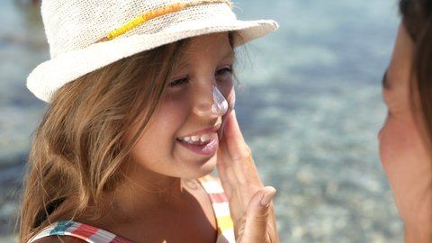 Close up of young happy mother applying protective sunscreen or sunblock lotion on her little happy smiling daughter's face to take care of skin on seaside beach during family holidays vacation trip.