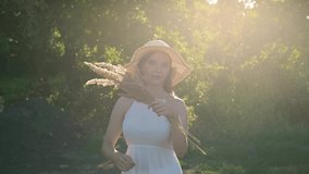 A girl in the rays of the setting sun is waving a bouquet of their dead wood. Grass particles in the air. Summer atmospheric video. Portrait of a beautiful girl in a brown hat