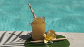 Video footage of glass mason jar with orange juice, bamboo straw, half of fresh orange, yellow frangipani flower, shade from palm tree and bubbling blue swimming pool on background.