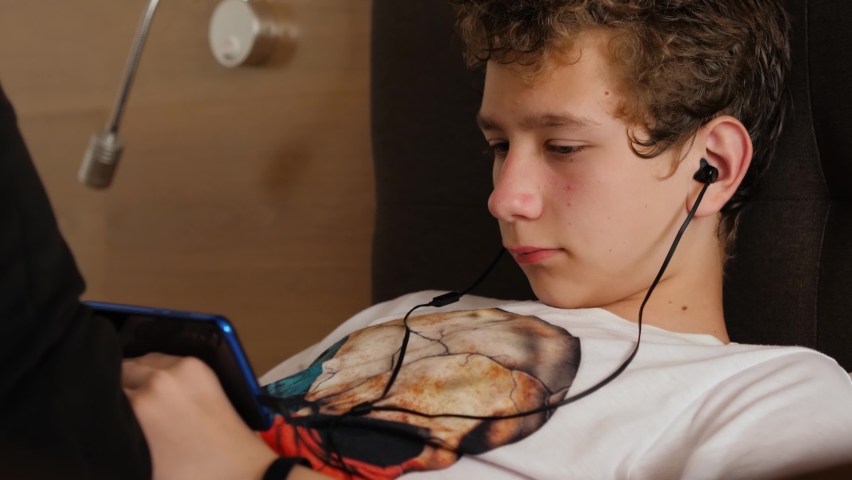 Young man is lying on the bed and looking for something in his smartphone. Teenage boy with headphones communicates on the Internet via a mobile phone Royalty-Free Stock Footage #1076767205