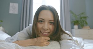 Close up of cheerful young Caucasian pretty woman in pajamas laying in bed at home and looking at camera smiling in good mood. Beautiful joyful female videochatting online in bedroom, rest concept