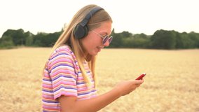 Slow motion video clip of pretty blonde girl teenager young woman wearing a striped t-shirt and blue sunglasses in a field listening to music on her cell phone and wireless headphones 