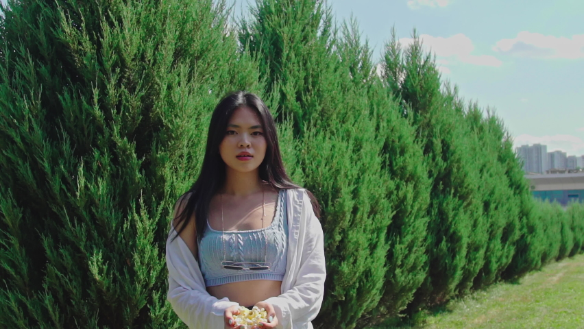 Girl scatters popcorn and looking at the camera over green trees background. Asian young woman in casual outfit outside. Attractive lady with dark hair and brown eyes sits on ton red stair. 4K