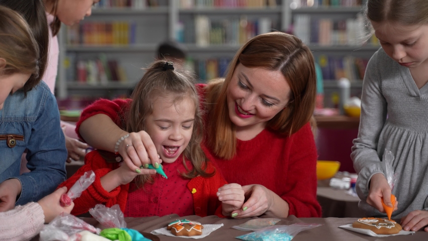 Close-up of teacher helping little girl with autism to decorate biscuit with glaze. Kids with disabilities decorating cookies with colorful icing during masterclass | Shutterstock HD Video #1076780711