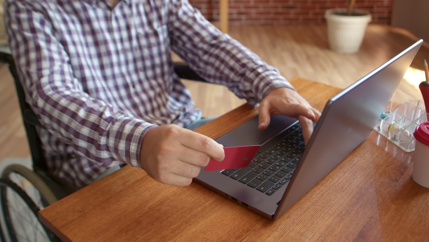 Disabled man sitting wheelchair front of laptop with bank card in his hand, filling out information, making purchase online. Concept of service availability