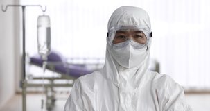 Video portrait shot of medical person wearing PPE, Personal Protective Equipment, clothes standing and looking to camera in hospital with saline solution and gurney in blur background.