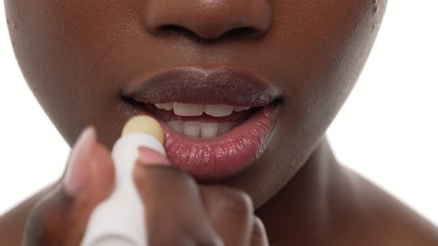 Close up of an african woman's mouth, applying lip balm on her top and bottom lips. White background.