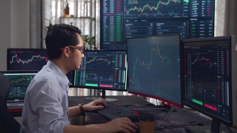 Asian Young Male Stock Market Broker Analysing Graphs On Multiple Computer Screens
