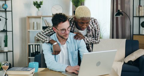 Homosexual relationship concept where cute smiling satisfied black-skinned guy with same sex orientation standing and putting his hand on positive joyful partner's shoulder which working on laptop at