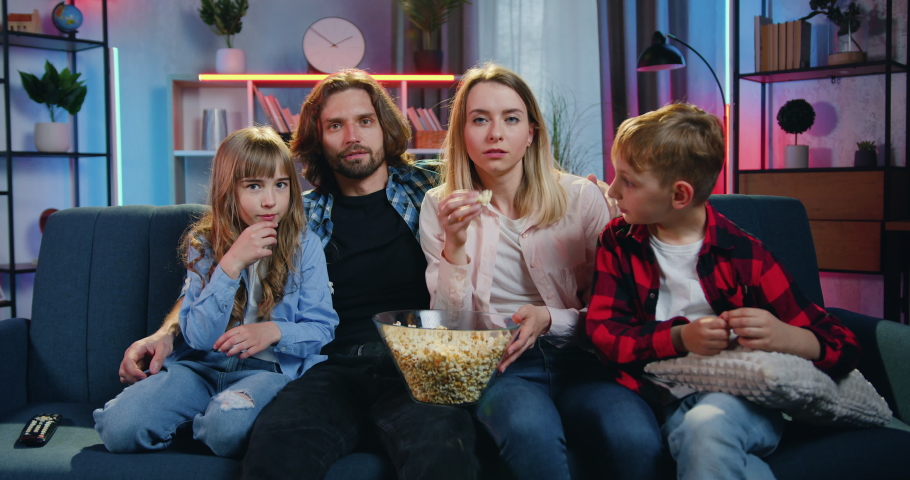 Portrait of lovely happy smiling friendly family of four persons-mother,father,son and daughter which sitting on the couch at home in the evening,watching TV and tasting popcorn | Shutterstock HD Video #1076815820