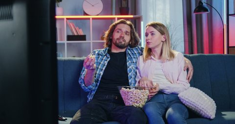 Family atmosphere concept where attractive carefree couple sitting on the couch in front of TV ,eating popcorn,talking and bouncing on the couch in fright when their two kids jumping out from behind