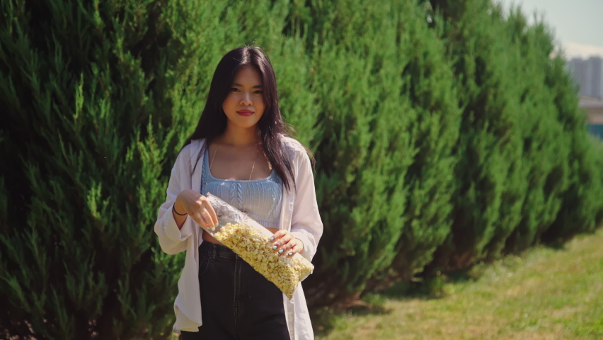 Girl eats popcorn looking at the camera in the park. Asian young woman in casual outfit outside. Attractive lady with dark hair and brown eyes