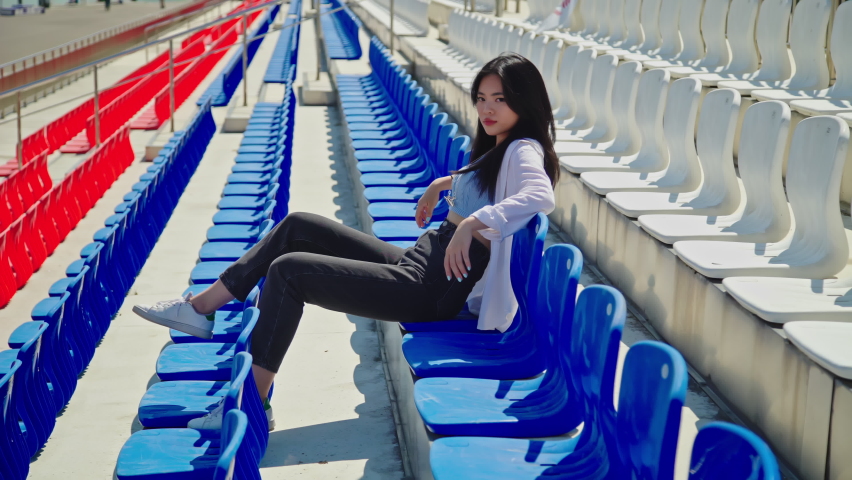 Beautiful young asian woman relaxes looking at the camera on tribune in hot day. Girl in casual outfit outside. Attractive lady with dark hair and brown eyes posing at stadium tribune 4K