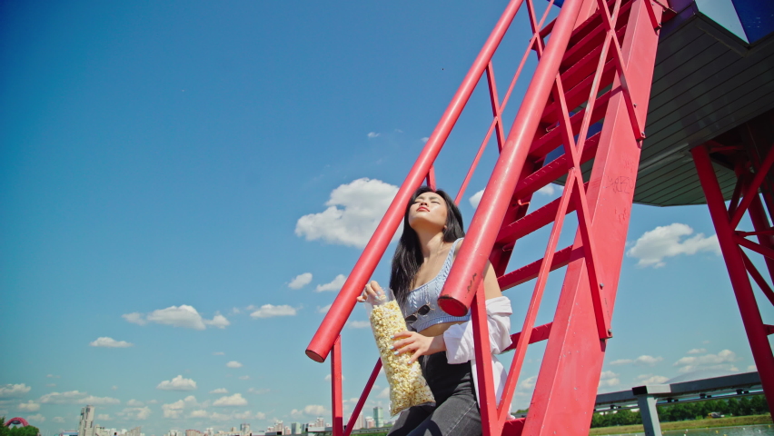 Beautiful young asian woman eating popcorn on red stair in the park on sunny day. Asian girl in casual outfit outside. Attractive lady with dark hair and brown eyes sits on ton red stair. 4K