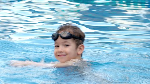 A preschool child in the pool in swimming goggles splashes on the water in the pool, a boy jumps and holds a swimming board in his hands.