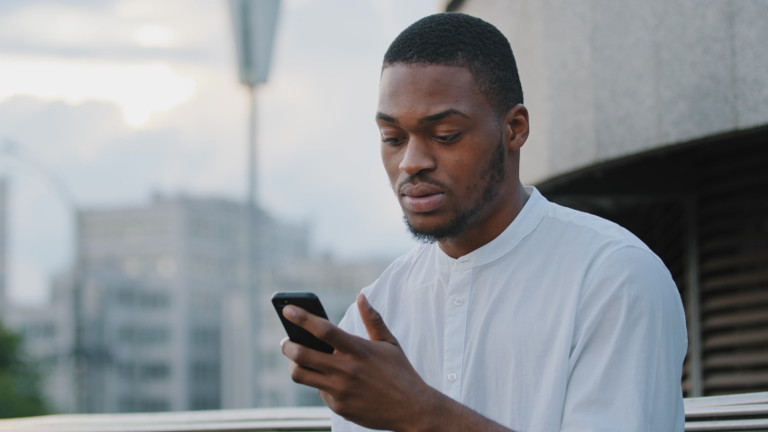 Male upset frustrated face afro american sad angry man african guy sitting on street outdoors feels irritated looking at phone checking late time in smartphone looking around waiting for someone | Shutterstock HD Video #1076817845