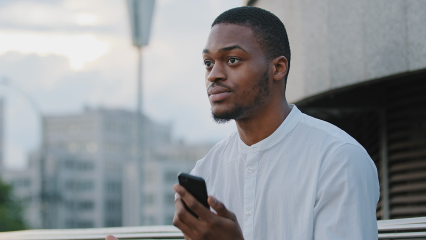 Male upset frustrated face afro american sad angry man african guy sitting on street outdoors feels irritated looking at phone checking late time in smartphone looking around waiting for someone | Shutterstock HD Video #1076817845