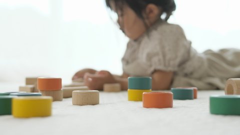 Toddler girl lying on a white floor mat and playing in a good relax mood with wooden building blocks at home in the living room. Slide motion
