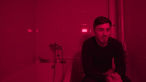 Cheerful Man Sits on the Toilet in a Nightclub, Shakes His Head in the Rhythm of Music and sings along. A Young Guy Sits In A Toilet In A Restroom Illuminated By A Red Lamp
