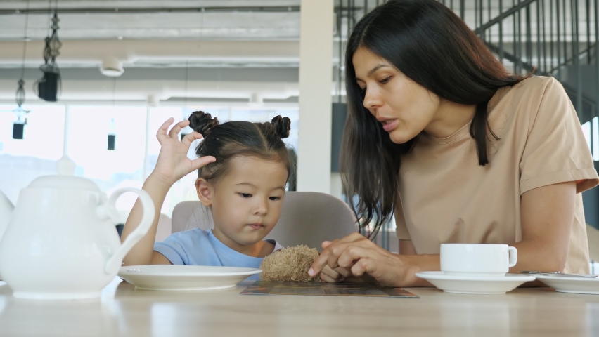 Mother with kid girl in restaurant. Asian young woman brunette and little toddler daughter choose menu sitting at brown wooden cafe table close view