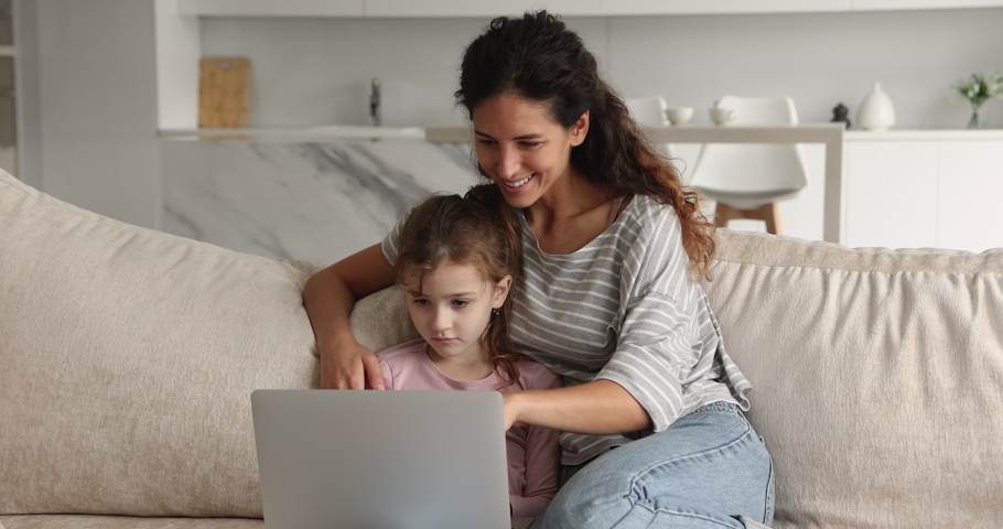 Affectionate caring young mother or nanny watching funny cartoons online on computer with little preteen kid daughter, resting together on comfortable couch at home, leisure weekend activity pastime. Royalty-Free Stock Footage #1076831066