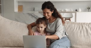 Affectionate caring young mother or nanny watching funny cartoons online on computer with little preteen kid daughter, resting together on comfortable couch at home, leisure weekend activity pastime.