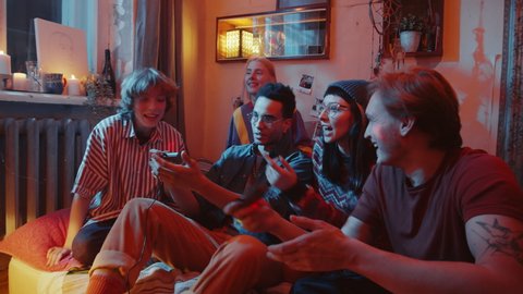 Company of young cheerful friends sitting together on bed in dark room with light projector, chatting and playing retro console game with excitement at home party