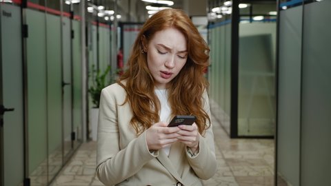 Worried businesswoman reading bad news in message on her smartphone while standing at office. Shocked and scared young female office manager holding mobile phone in hands indoors