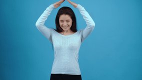 The charismatic, optimistic girl raises her hands up like a roof over her head. Asian with dark hair, dressed in a blue blouse, isolated on a dark blue background in the studio. The lifestyle concept