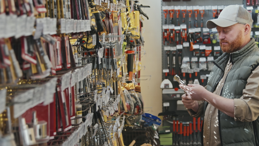 Slowmo PAN of bearded man in puffy vest and cap looking at wrench at hardware store, then turning towards camera Royalty-Free Stock Footage #1076840654
