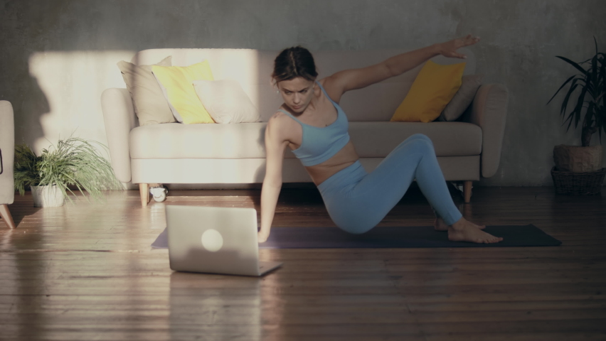 Adult caucasian woman practicing on a yoga mat, using her laptop for an online yoga class instructions. Doing yoga at home in sunlight beams.