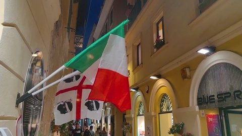 Alghero, Sardinia, Italy - 07 28 2021: Two flags of Italy and Sardinia island fluttering side by side in a narrow old town street of Alghero.