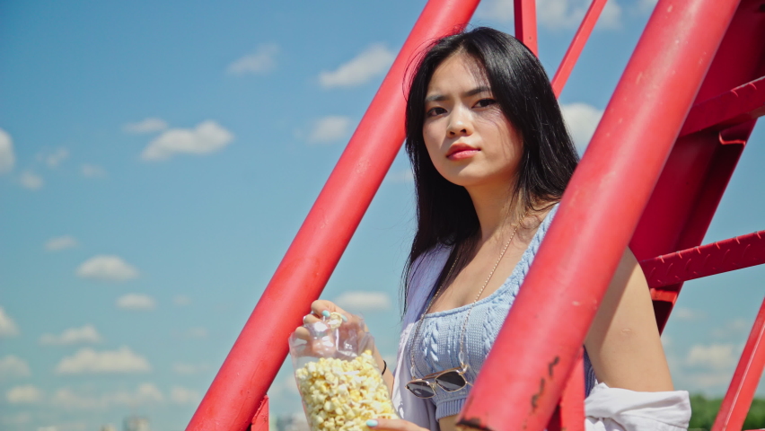 Beautiful young asian woman eating popcorn while sitting on the red stairs in the park on a sunny day. Girl in casual outfit outside. Attractive lady with dark hair and brown eyes 4K