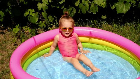 A little girl splashes in the water in a small inflatable pool in the garden. A hot sunny day with water activities. Children's portable mini pool
