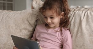Happy adorable small child girl holding digital computer tablet in hands, playing online games, having fun communicating distantly in social network or web surfing, modern technology addiction concept