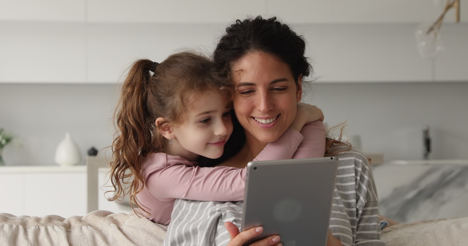 Loving small preteen kid girl embracing beautiful young mommy or nanny, using digital computer tablet together at home, web surfing information, playing games or shopping in internet store together. Royalty-Free Stock Footage #1076855726
