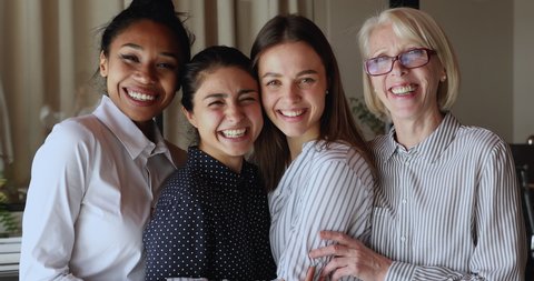 Happy beautiful multiracial multi generational independent women in formal wear standing indoor hugging laughing. Four diverse company staff members friends enjoy good relations and friendship at work