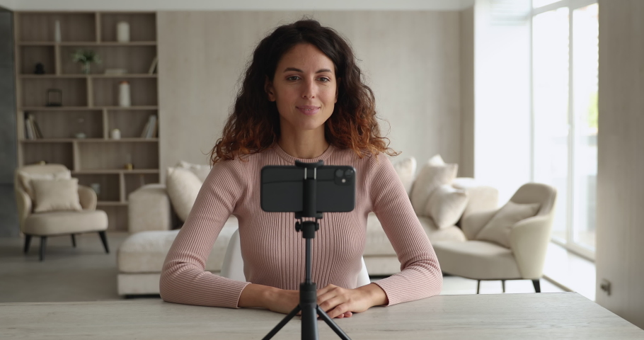 Smiling confident young hispanic female blogger sitting at table in front of cellphone on stabilizer, talking speaking recording video or streaming online in social network, virtual event concept. Royalty-Free Stock Footage #1076855864