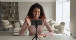 Smiling confident young hispanic female blogger sitting at table in front of cellphone on stabilizer, talking speaking recording video or streaming online in social network, virtual event concept.