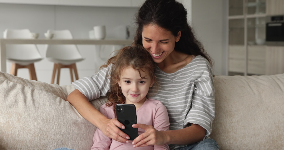 Joyful young nanny playing mobile games with adorable small child girl, resting on comfortable sofa in living room. Addicted to modern technology happy mum and kid daughter having fun using cellphone. Royalty-Free Stock Footage #1076855972