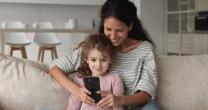 Joyful young nanny playing mobile games with adorable small child girl, resting on comfortable sofa in living room. Addicted to modern technology happy mum and kid daughter having fun using cellphone.