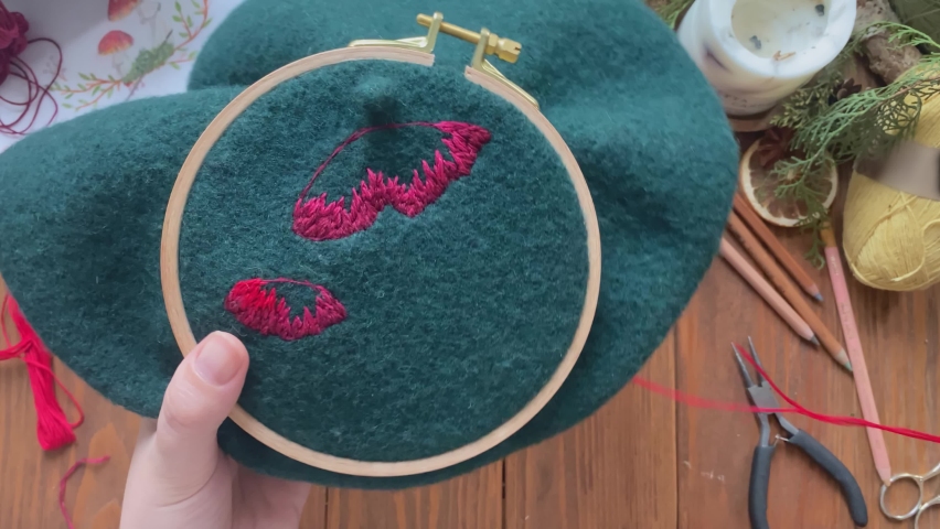 Embroidery process with red cotton thread of mushrooms hat. Needlework concept. Woman's hands sewing fly agaric on green beret. Craft and handmade. Wooden table with yarn scissors pencil and candle. | Shutterstock HD Video #1076860484