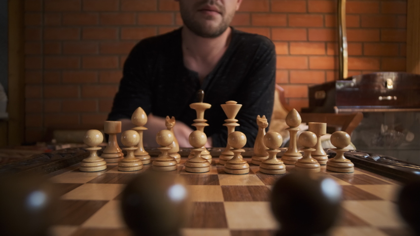 unrecognizable young bearded man in black T-shirt makes move with wooden pawn on chessboard against background of red brick wall.