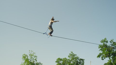 SLOW MOTION: Man slacking on highline against the sky above the big urban city on a beautiful summer day