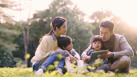 asian family with two children sitting on grass talking relaxing outdoors on in park at sunset